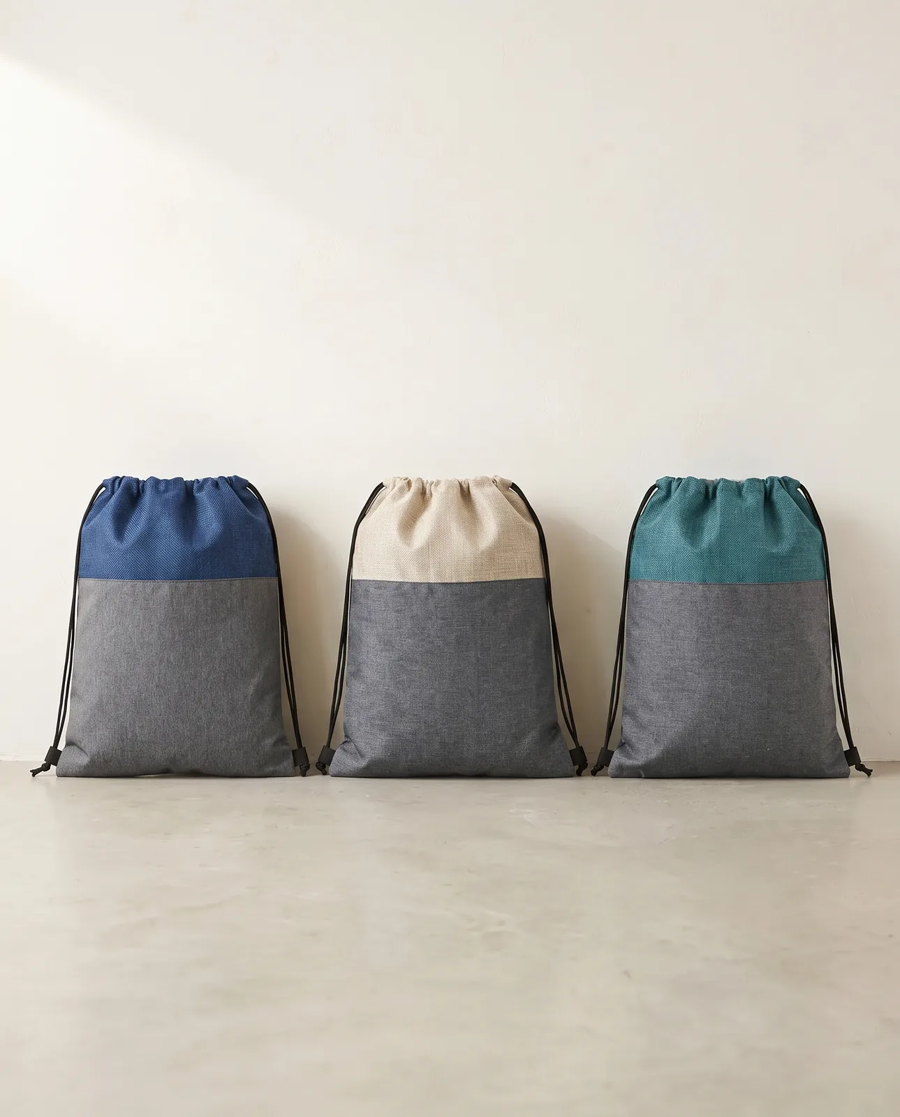 Custom cinch backpacks in Blue, Natural Beige and Teal Blue lined up on a concrete floor