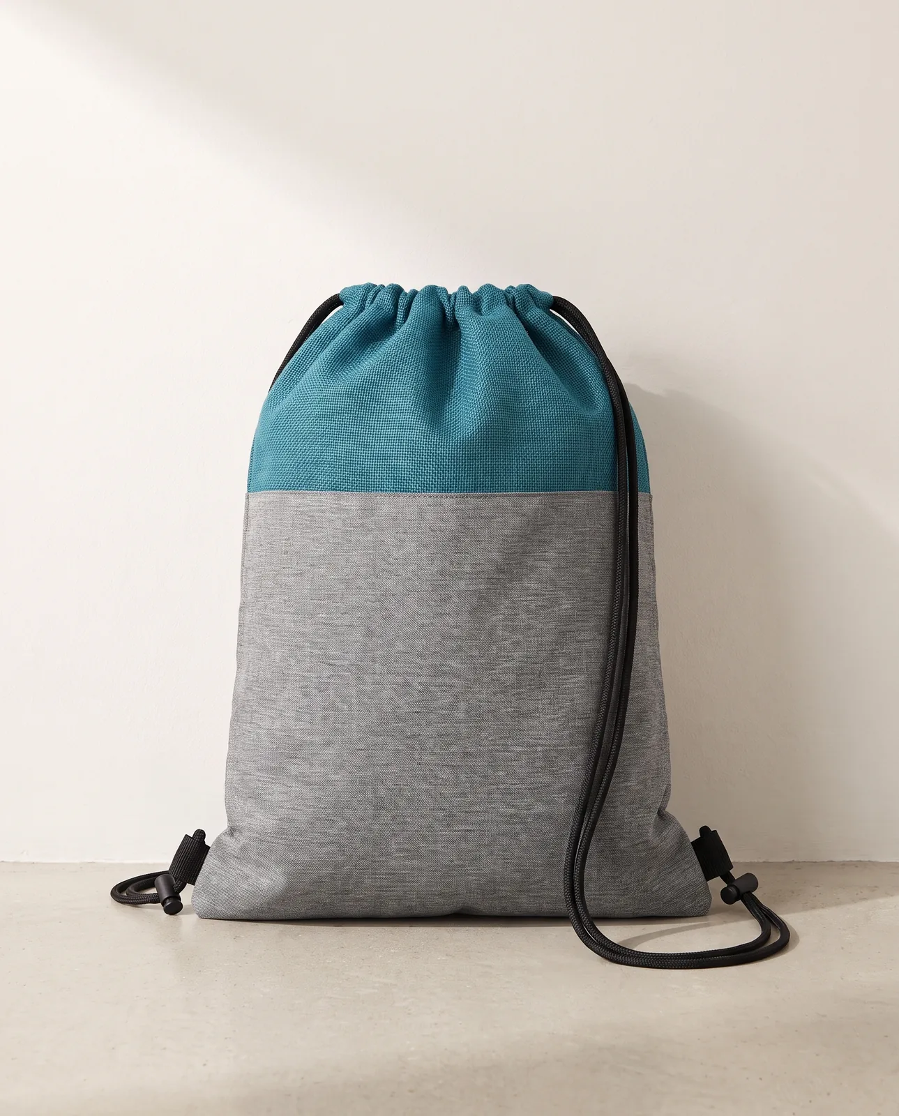 Custom cinch backpacks in Teal Blue, heathered grey body with a teal jute-look top panel and black cords, standing against a white wall