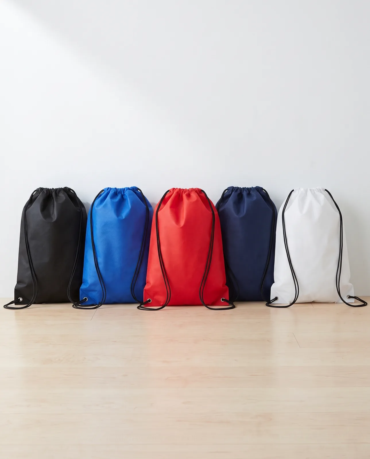 Custom drawstring bags in Black, Royal Blue, Red, Navy Blue and White lined up against a white wall