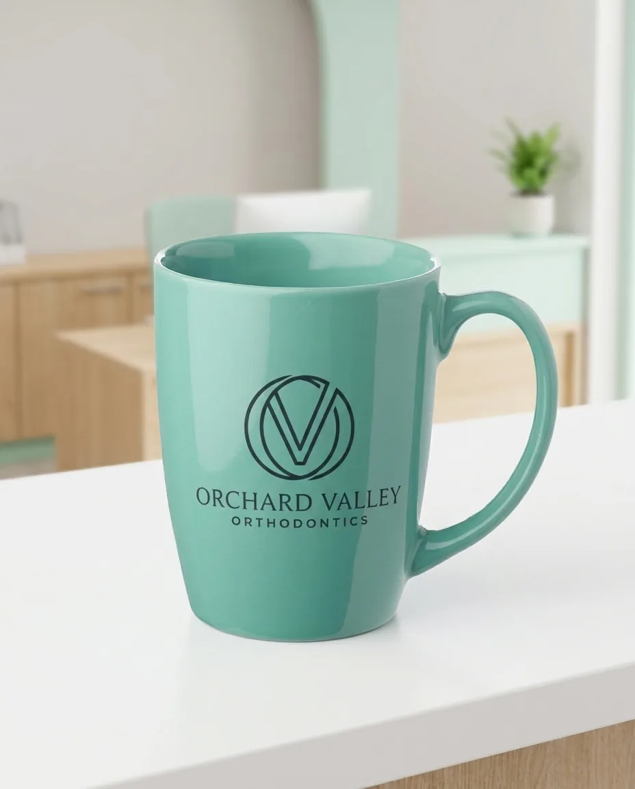 Seafoam teal promotional coffee mug with its high curved handle turned to the camera on a dental clinic counter, with a one color orthodontics monogram screen printed on the front