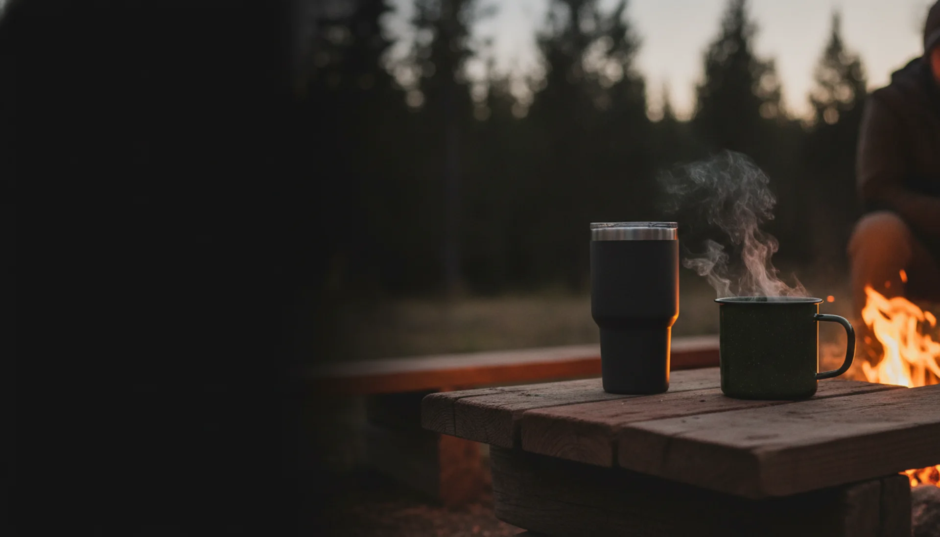 Custom insulated tumbler and enamel camp mug beside a campfire at dusk