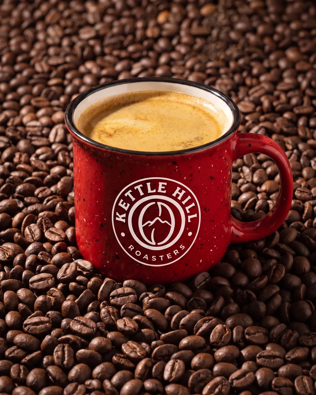 Red speckled 7.5 oz custom campfire mug with a one color white roaster badge, holding a double espresso on a bed of coffee beans