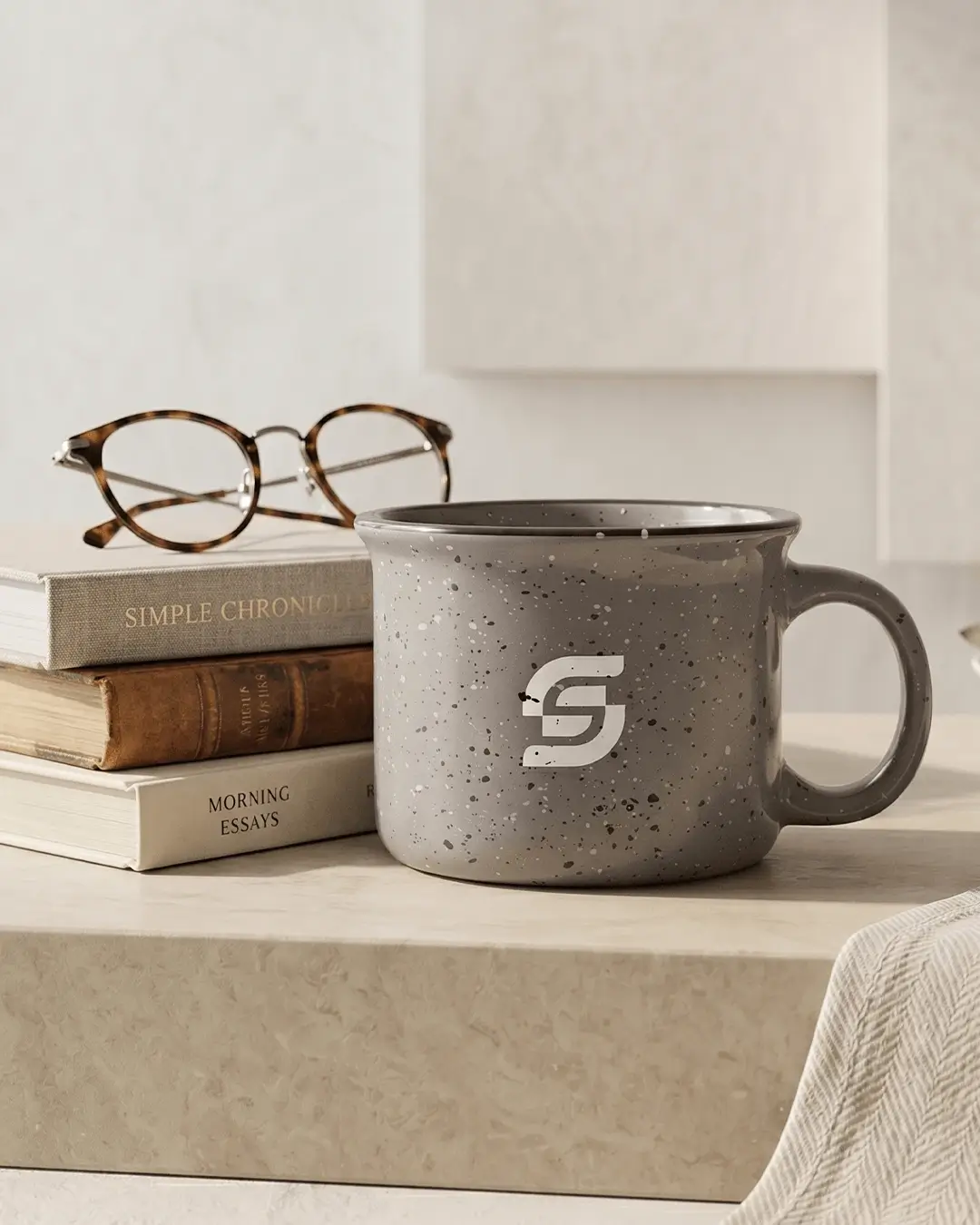 Gray speckled custom campfire mug with a printed logo beside a stack of books and glasses