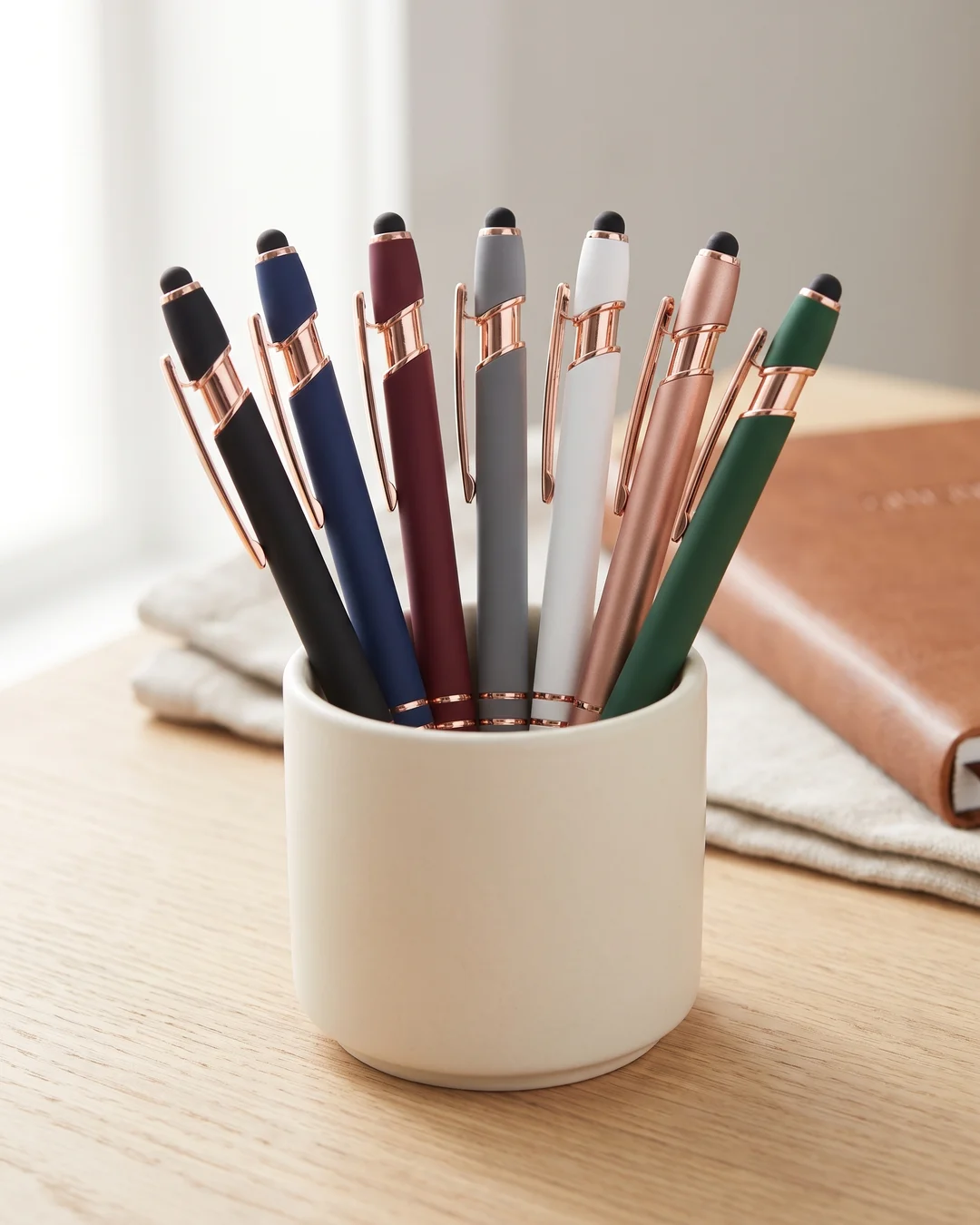 Custom engraved pens in several soft-touch colors with rose gold trim standing in a ceramic cup on a hotel front desk