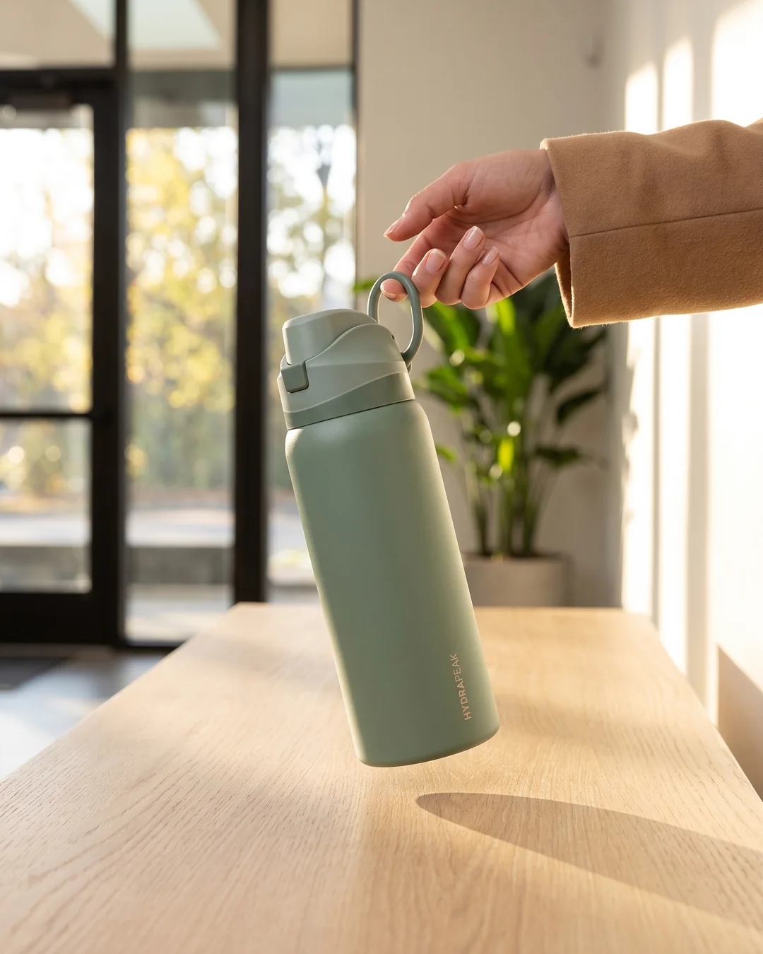 Hand carrying a Hydrapeak 32 oz Oasis water bottle in Sage Leaf by its loop handle in a bright office entryway