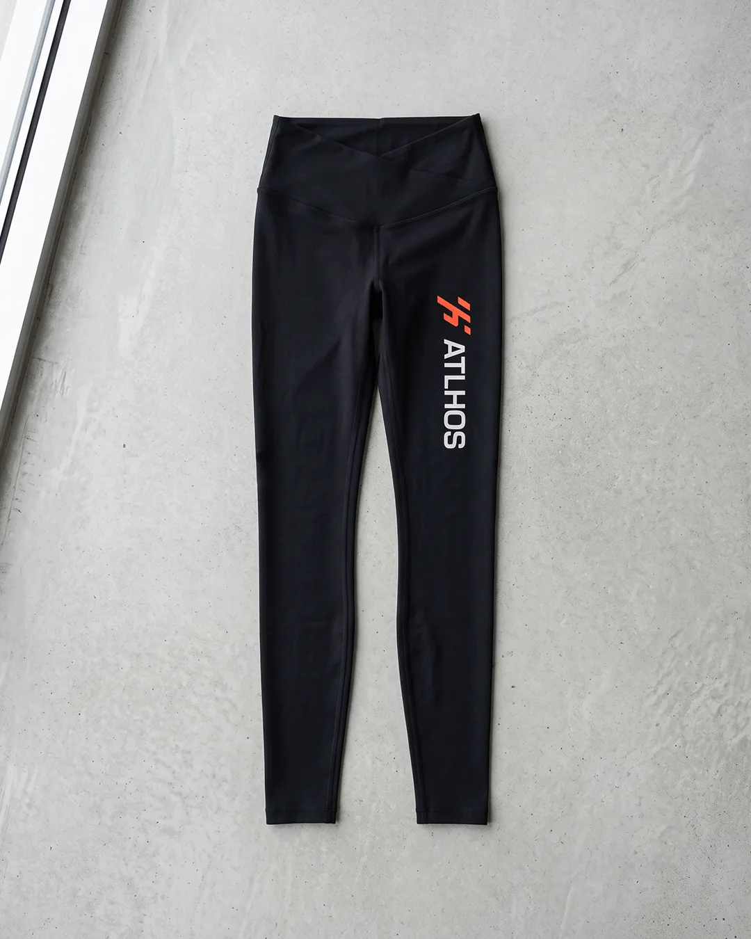 Custom leggings in Black laid flat on concrete, showing the crossover V-front waistband and a two color logo printed down the upper left leg