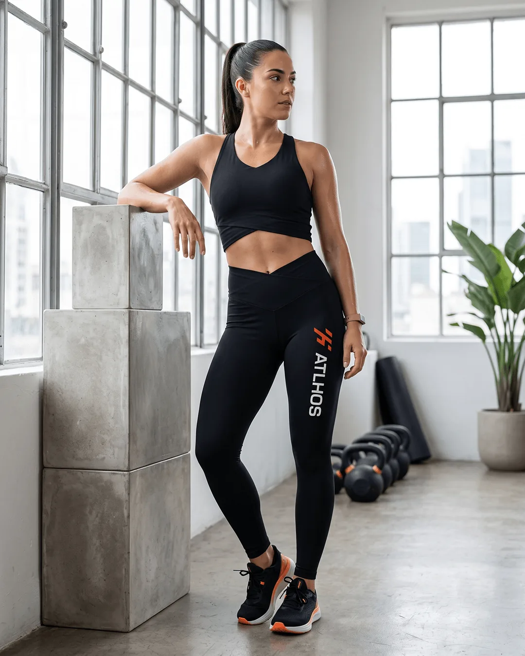 Woman in a bright gym wearing custom high-rise leggings in Black with a crossover waistband and a printed logo running down the left leg