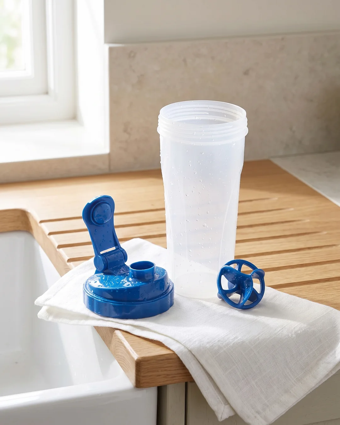 Custom shaker bottle taken apart after washing: the clear cup, the blue flip-top lid and the blue Vortex mixing ball on a towel by the sink